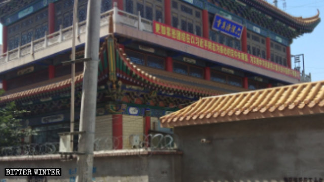 The wall surrounding a mosque is covered with barbed wire, a propaganda slogan displayed above the entrance.